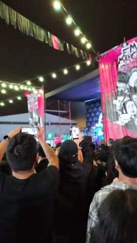 A concert is in progress with a band performing on stage, which is decorated with pink banners featuring the text "EMO MERCHANT". The crowd is gathered, with many individuals holding up their phones to record the performance. The stage is lit with blue, green, and purple lights. The environment appears to be an outdoor venue, partially covered. There are string lights hanging overhead and scaffolding visible on the side of the stage.