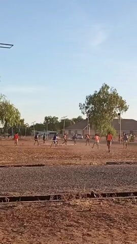 A video shows a dusty, dirt field with several people playing soccer in the distance. The field appears to be under construction, as there are piles of gravel and recently installed solar-powered streetlights lining the area.  Several children are running and playing soccer in the distance. The sky is clear and blue. A residential building is visible in the background. The camera is fairly static and slightly shaky, seemingly held by hand. A plastic bag sits in the lower right corner.