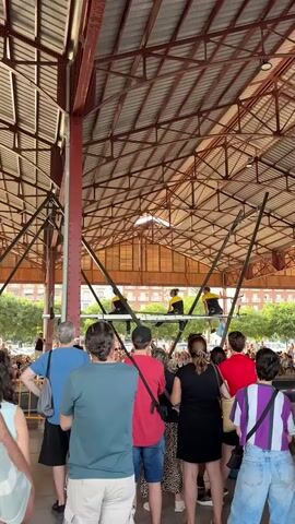 A group of four performers, each wearing a yellow shirt and black pants, are positioned on a horizontal bar suspended by cables. They appear to be engaged in a balancing act, with one performer standing and the others seated or in various crouching positions. The performers are at a considerable height, likely for an audience. Below them, a diverse crowd of people stands watching the performance. The environment is an open-air structure with a visible roof and supporting beams, suggesting a market or public gathering space. The people in the foreground are facing away from the camera, engrossed in the show. The day appears to be bright and sunny, with trees and buildings visible in the background outside the structure.
