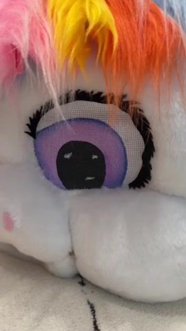 This video shows a close-up view of a stuffed unicorn head. The unicorn has a white plush exterior, with a rainbow-colored mane made of fluffy yarn in pink, orange, yellow, blue, and purple. Its eyes are large and circular with black pupils and a purple and pink iris. The eyelashes are thick and black. A shimmering purple unicorn horn with spiral detailing is visible at the top of its head. The background is slightly blurred, but appears to be a textured surface, possibly a blanket or rug. The lighting is bright and appears to be coming from overhead. There are no people or other discernible objects in the frame. The video is short and static, with no movement or sound.