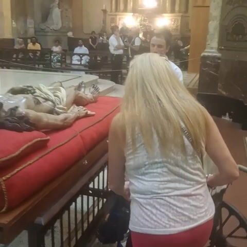 The scene takes place inside a church or cathedral. A life-sized statue of the deceased Jesus Christ is laid out on a red velvet-covered platform. A woman with long blonde hair, wearing a white patterned top and red pants, stands facing the statue, while an older woman with blonde hair, seated in a wheelchair, reaches out to touch the statue. A man in a white robe stands nearby, observing. Other people can be seen sitting in the background, some on benches. The floor is tiled, and there are religious artifacts and artwork visible in the surroundings.
