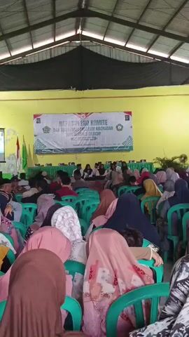 A large group of people are seated in a covered outdoor area, facing a stage. The people appear to be mostly women and children, many wearing hijabs. The stage has a banner with text and logos, and there are flags on either side. The environment appears to be a community gathering or event.