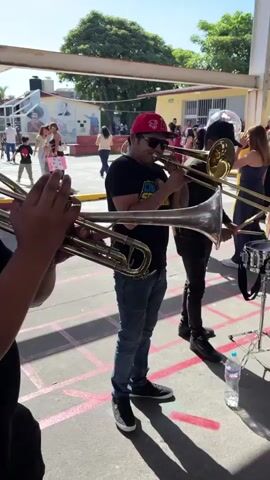A group of people are playing musical instruments outdoors. In the foreground, a person wearing sunglasses and a black shirt plays a trumpet. To their right, another person in a red cap and black shirt plays a trombone. Further to the right, a person in a black shirt and jeans plays a snare drum. Another person in a black shirt and jeans plays a bass drum with "BBS" written on it. In the background, there are other people, some of whom appear to be watching the musicians. There are also balloons and decorations visible, suggesting a celebratory event. The scene appears to be in a courtyard or open area with a roof structure overhead.
