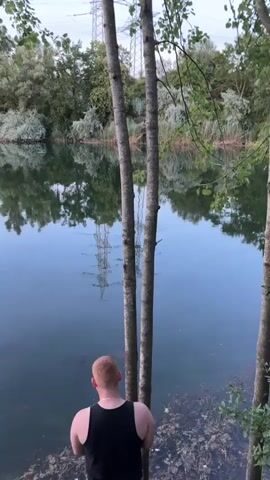 A man with short red hair wearing a black tank top stands at the edge of a body of water. He is leaning against two thin trees. The water reflects the trees and the surrounding greenery, as well as a power line tower in the distance. The sky above is overcast with some visible power lines stretching across it. The ground at the edge of the water is covered in leaves and debris. The camera pans slowly to the right, showing more of the lake and the trees on the opposite bank.