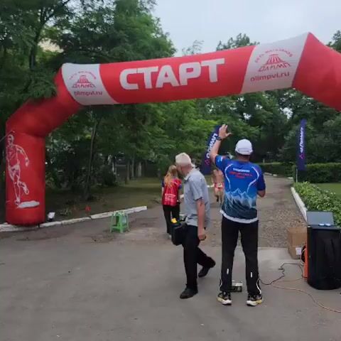 A group of people are gathered near an inflatable archway with the word "START" written on it in large white letters. The arch is red and white, and has logos on it. Two men and a woman are standing in the foreground. The woman is wearing a red vest. The man on the right is wearing a white cap and a blue and white shirt. The man on the left is wearing a grey shirt and black pants. A runner, wearing orange shorts and no shirt, is approaching the archway. Another person is visible in the background, running. The environment appears to be outdoors, with trees and foliage visible in the background. There are also some small green stools placed on the ground. A black speaker-like object is visible on the right side of the frame.