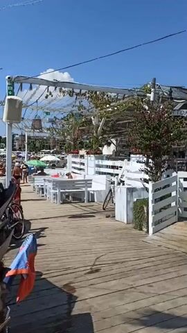 The video pans across a sunny outdoor cafe on a wooden boardwalk. White tables and benches are set up under a white canopy adorned with fairy lights and greenery. To the right, a bar area is visible with bottles of alcohol and glasses. Several people are present: some are sitting at tables, others are walking along the boardwalk, and a few are at the bar. A woman in a black dress is seen standing near a small food kiosk, and a man with a backpack is walking past. In the background, beach umbrellas and people on the beach can be seen. The overall atmosphere is one of a relaxed beachside establishment on a clear, bright day.