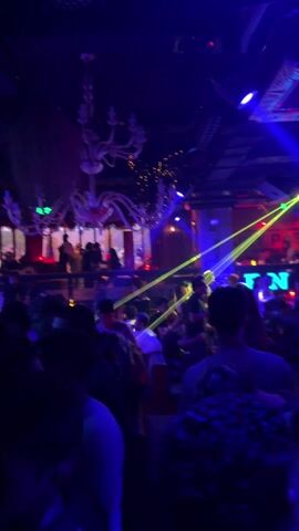 The video shows a crowded nightclub scene. Many people are standing close together, likely dancing. Laser lights and spotlights in green and blue illuminate the space from the ceiling, where there is a chandelier. The club has a dark, industrial feel with exposed beams and other fixtures on the ceiling.