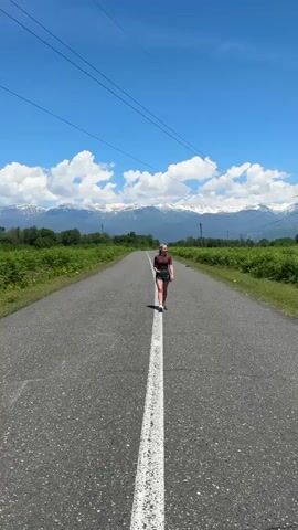 A young woman is walking towards the camera on a long, straight asphalt road. She is positioned in the center of the road, where a solid white line runs down the middle. She wears a dark maroon short-sleeved top, dark shorts, white sneakers, and a light-colored baseball cap worn backward. Her expression is relaxed, with a slight smile.

The road is flanked on both sides by lush green foliage and trees, indicating a natural or rural setting. In the distant background, a majestic mountain range with snow-capped peaks stretches across the horizon under a bright blue sky dotted with fluffy white clouds. Power lines are visible overhead, supported by utility poles along the road. The weather appears sunny and clear. The scene depicts a serene and picturesque landscape in Akwaskia tshyts, Abkhazia.