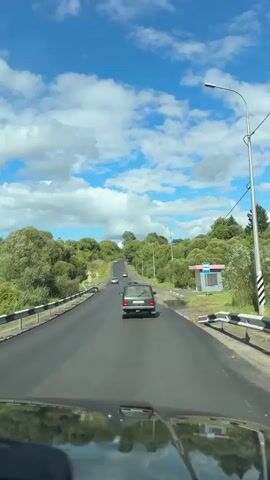 A dash cam view from inside a vehicle shows a paved road with a few cars ahead. The road is surrounded by green trees and vegetation on both sides, with a bright blue sky dotted with white clouds above. A bus stop shelter with a blue roof and sign is visible on the right side of the road. The vehicle is driving uphill, following the curve of the road. The dashboard of the camera vehicle is reflected in the windshield.