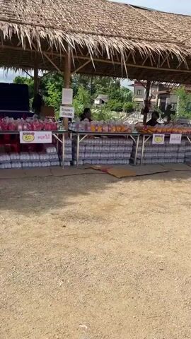 The video shows an outdoor market stall under a thatched roof. Several tables are laden with goods. On the left, there are stacks of bottled water and several bags of what appear to be snacks or food items. In the center and to the right, more tables display bags of goods, some with price tags indicating 60 and 50 Thai Baht. Two women are visible behind the tables, one appears to be arranging items. Further to the right, two men are seated at tables with what look like game boards or displays. A young man wearing a blue shirt and a backpack walks past the stall. In the background, there is lush greenery and a white and gold structure that resembles a shrine or monument, complete with a dragon sculpture. The ground is dirt and gravel. The sky is cloudy.
