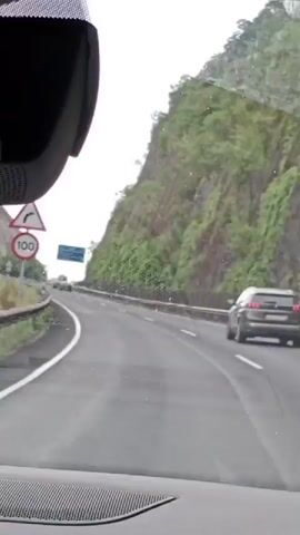 This video shows a view from inside a car driving on a highway. The car is moving along a road that is bordered by steep, rocky hillsides covered in vegetation. The sky is overcast. There are other vehicles on the highway, including a dark-colored SUV in the right lane and at least one car further ahead in the left lane. A blue highway sign is visible in the distance. The camera is positioned to capture the road ahead and the surrounding scenery through the windshield.