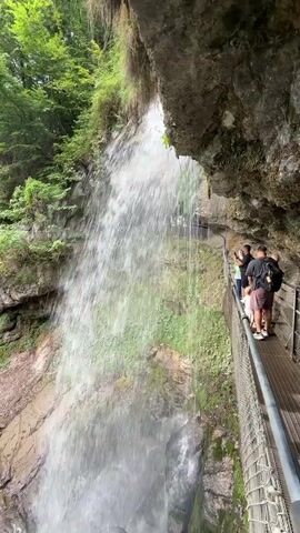 The video shows a scenic view of a waterfall cascading down a rocky cliff face. A narrow metal walkway with a mesh railing runs along the side of the cliff, allowing people to walk behind the waterfall. Several individuals are visible on the walkway, observing the waterfall. Beyond the waterfall, lush green trees cover the surrounding hills. Further in the distance, a large, multi-story building with a red roof is visible amidst the trees, with a body of turquoise water to its left and a mountain range in the background. The water flows into a rocky riverbed below the waterfall, with a few fallen logs scattered in the water. The overall scene depicts a natural landscape with man-made elements for visitor access.