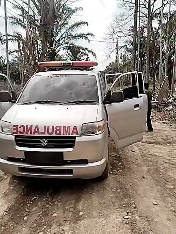 The video shows a silver ambulance parked on a dirt road in a rural area. The driver's side door is open. Several people are standing in the road near the ambulance. Some of them are wearing headscarves. There are trees in the background, and a small house can be seen in the distance.