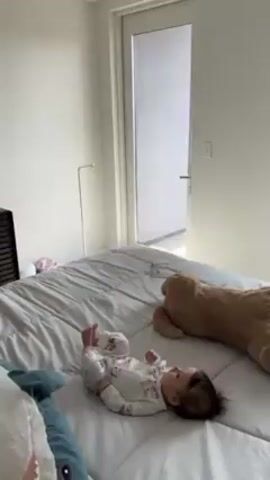 The video begins with a baby lying on a bed next to a stuffed bear. The baby is wearing white pajamas with a small print. The bed has a gray cover on it, and there are throw pillows at the edge of the bed. The camera then pans upward to the ceiling, showing a ceiling fan with dark wooden blades and a white light fixture in the center. The ceiling and walls are white. There is a white door in the background.