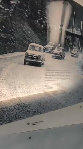 The video pans across a museum exhibit featuring large, black-and-white photographs mounted on a curved wall. The photographs depict historical scenes, possibly related to transportation or infrastructure development in a mountainous region. Several cars are visible in some of the images, along with groups of people, and what appear to be construction or maintenance activities. One section of the exhibit is labeled "Eröffnung," suggesting an opening ceremony. Another section shows images of winter conditions. The exhibit is well-lit, with some spotlighting highlighting specific areas. A man in casual clothing is briefly visible standing and observing the exhibit. The overall environment appears to be a modern museum space with a textured ceiling and polished floor.