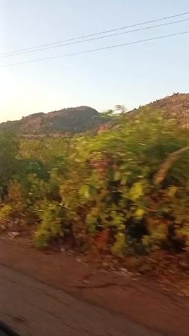 The video shows a view from a moving vehicle. The camera is positioned near the window, showing the passing scenery. The landscape consists of hills covered with green vegetation and some dry grass. The ground near the road is reddish brown. Several power lines and at least one utility pole are visible against a clear, light blue sky. The overall impression is that the vehicle is traveling through a rural, somewhat hilly area.