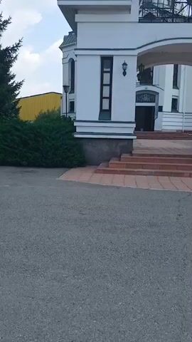 The video begins with a low-angle shot of the front of a white building with a green roof. The building has an arched entranceway with a set of brick stairs leading up to it. A smaller arched section is visible above the main entrance, containing decorative metalwork and a large stained-glass window. A golden dome with a cross on top is visible at the peak of the roof. The sky is blue with some white clouds. To the left of the building, a dark green bush is visible, and behind it, a yellow building with a corrugated metal roof can be seen. The video pans slowly upwards, revealing more of the building's facade, including windows and decorative light fixtures. A blue sign with white text is attached to the building near the right side of the entrance. The video then continues to pan upwards, focusing on the dome and the sky.