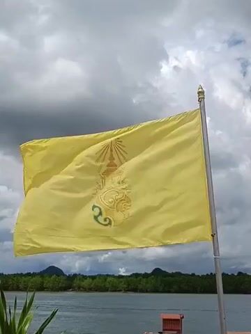 A yellow flag with a royal crest is fluttering in the wind, set against a cloudy sky. Below the flag, a calm body of water is visible, with a distant treeline and hills on the horizon. In the foreground, part of a boat or pier can be seen. The video appears to be taken from a stationary viewpoint.