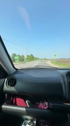 The video shows a view from inside a car driving down a straight, paved road under a clear blue sky. The dashboard of the car is visible in the foreground, with an air vent on the left and a storage compartment below containing a red cloth and some pink tissues. The road has white lines marking the lanes and appears to be in a rural area with trees and fields on either side. There are lampposts and a pedestrian crossing sign visible along the road. At one point, a dark-colored SUV passes the car. The video is shot from the passenger's perspective, looking out through the windshield.