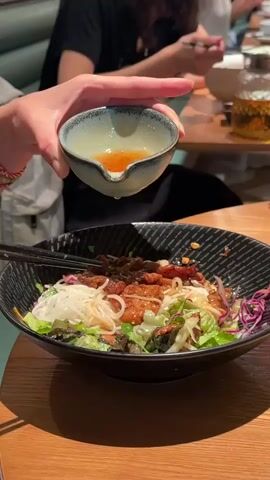 A person's hands are shown holding a small, light blue bowl with a dark rim over a dark, patterned bowl containing a noodle dish. The person is pouring a light brown liquid from the small bowl into the noodle dish. The noodle dish appears to contain white rice noodles, green leafy vegetables, red cabbage, and pieces of meat, possibly chicken or pork. There are black chopsticks resting on the edge of the noodle dish. In the background, other people are seated at a wooden table, and a glass teapot with a golden liquid is visible. The overall environment appears to be a restaurant setting.