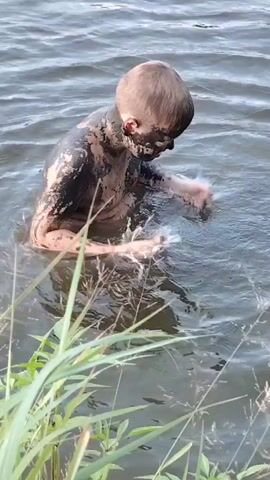 A young person covered in mud is standing in murky water. They are wiping mud from their face with their hands, splashing water. The water is still except for the disturbance caused by their actions. Green reeds are visible in the foreground. The person’s eyes are squeezed shut and their mouth is open in a grimace.