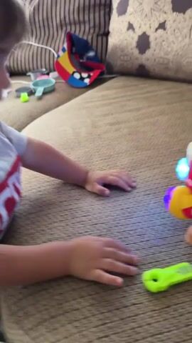 A young child lies on a couch, playing with toys. The child's hands are visible as they interact with a french fry-shaped toy that has animated eyes and a mouth. A toy knife with a green handle lies nearby. The child leans in to touch the french fry toy, and a red light appears to emanate from it. The child's t-shirt has a red stripe with white lettering across the chest. Various other toys are scattered on the couch in the background.