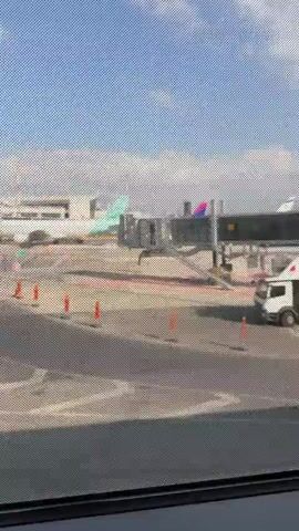 The video shows an airport tarmac viewed through a window. In the distance, an airplane with teal and white livery is parked at a gate, connected to a jet bridge. Various airport vehicles and equipment, including a white truck, are visible on the tarmac. Orange traffic cones mark a pathway. The sky is blue with scattered clouds. The window has a mesh pattern that slightly obscures the view. The camera pans slowly to the right.