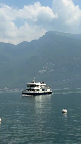 A white ferry boat with a blue stripe travels across calm, dark blue water. The ferry has multiple decks with people visible on them. Two white buoys are floating in the water. In the background, hazy, green mountains rise under a cloudy, pale blue sky.