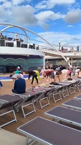 On the wooden deck of a cruise ship, a group of people of various ages engage in a stretching exercise, guided by a seated instructor. A line of unoccupied deck chairs sits in the foreground. The ship's superstructure and an open railing are visible in the background against a blue sky with scattered clouds.