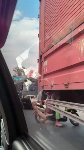 The video shows a traffic jam from the perspective of someone inside a car. A large red container truck dominates the right side of the frame, its side panel showing the letters "KT" and the number "22". To the left of the truck, other vehicles are visible, including a smaller truck with a tarp-covered load. A motorcycle is parked or moving slowly on the right side of the road, partially obscured by the truck. The road surface is visible, and the sky above is cloudy. The overall impression is of congested urban or semi-urban traffic.