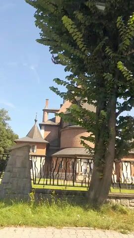 A video displays a tree in the foreground with a building in the background. The building appears to be a church or a similar structure with brick walls and a distinctive architectural style. There is a fence in front of the building, and the area around it is covered in grass. The sky is blue with a few clouds, suggesting it is daytime. The video has a timestamp of 00:00 to 00:04.