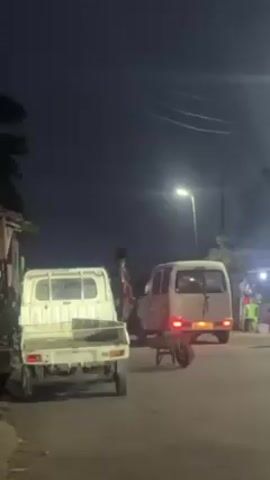 The video depicts an outdoor nighttime scene in what appears to be a residential or commercial area. Two small white trucks are parked on the side of the street, one of which is a van with its taillights on. A wheelbarrow is positioned between the two vehicles. People are visible walking around, and a small shop or kiosk is illuminated in the background with several people seated nearby. Streetlights provide the primary source of illumination. The overall impression is of a quiet, dimly lit street scene at night.