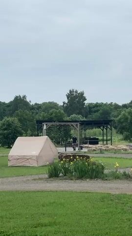The video pans across a campsite with several large canvas tents set up on a grassy field. In the background, there is a covered outdoor structure with people standing underneath it. Further back, more tents and some yellow buildings are visible, along with a white SUV parked on a gravel path. The landscape is dotted with trees, and the sky is overcast. A large bale of hay sits in the foreground, flanked by two red posts.