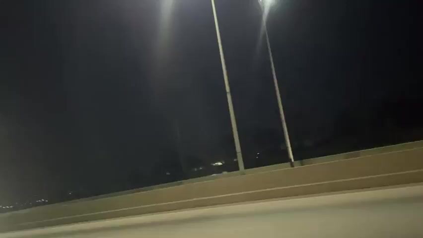 The video captures a nighttime scene on a highway, likely in Nigeria, based on the context provided. The road is illuminated by a series of tall streetlights, casting a bright glow on the asphalt. Several vehicles are visible, including what appears to be an ambulance with flashing red and blue lights, and a white bus. The overall atmosphere is that of a moving vehicle on a journey.

The activity taking place is travel on a highway at night. The presence of the ambulance suggests a potential emergency situation or a response to one. The streetlights indicate urban or semi-urban infrastructure. The darkness of the sky points to it being late at night or early morning. There are no discernible people or animals in the frame, and the focus is primarily on the road and the vehicles. The quality of the video is somewhat grainy, as is common with low-light conditions. No text is visible in the video.