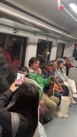 A man in a green jacket and khaki pants sits in a train car holding a microphone and a violin, apparently performing for the other passengers. He's surrounded by other people seated in the train. One person is recording him with their phone. Other passengers seem to be casually watching. The interior of the train is clean and modern-looking, with large windows showing blurry reflections of the outside.