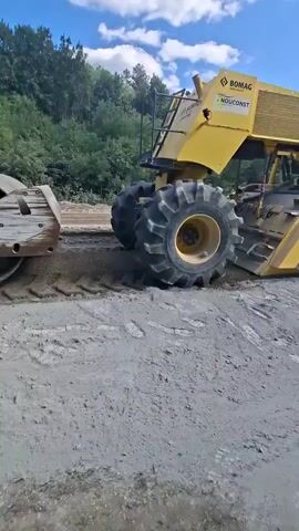 A large yellow Bomag milling machine is operating on a dirt road, grinding up the surface. The machine has large tractor tires and a milling drum at the front. The milling drum has the text "FLEX MIX" on it. The machine is moving from left to right in the frame, and the ground is being churned up. The sky is blue with some clouds, and there are trees in the background.