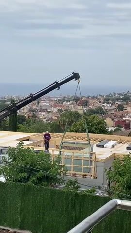A construction worker stands on a rooftop as a crane lowers a large glass structure. The structure is enclosed in a wooden frame and is suspended by green straps attached to the crane's hook. The rooftop is under construction, with exposed wooden beams and some covered materials. The background shows a city with red-tiled roofs and a distant body of water under a cloudy sky. Green foliage is visible in the foreground and on the side of the building.