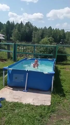 A boy is splashing in a small, blue, above-ground swimming pool. The pool is located on a concrete pad surrounded by green grass and a wooden fence. Behind the fence, there are trees and a bright blue sky with some clouds. The boy is wearing dark-colored swimming trunks and is jumping up and down in the water, creating splashes. The camera is positioned at a slightly elevated angle, looking down at the pool and the boy. The lighting suggests it is daytime and sunny.