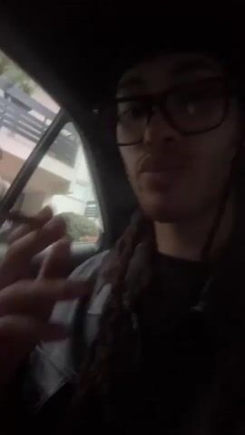 A person is smoking in the back seat of a car. The camera is pointing from the person's perspective out the car window toward another person standing outside. There is a can of alcohol sitting on the car door near the handle. The video then shifts so it is looking through the window, and the building is blurred due to movement.