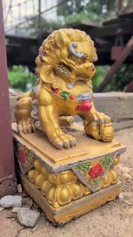 A close-up shot focuses on a golden lion statue, commonly known as a Foo dog or guardian lion, which is positioned on a decorative base. The statue is made of a metallic material, likely painted gold, and features intricate details on its mane, face, and body. The base it rests on is also gold, adorned with ornate patterns, including what appear to be red floral motifs and green leafy designs. The statue is placed outdoors on a ground surface composed of dirt, small stones, and debris. In the background, a blurred industrial or construction site is visible, with metal structures and greenery. The lighting suggests it is daytime, and the overall scene is somewhat rustic and aged, with dust or dirt visible on the statue and its base.