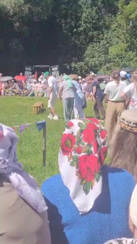 A group of people are participating in a tug-of-war competition on a grassy field. The participants are gathered around a rope, pulling on it with effort. Spectators are seated and standing in the background, observing the event. The environment appears to be outdoors, with trees and sunlight suggesting a warm, possibly summer day. The people in the foreground are wearing headscarves.