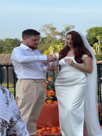 In a bright outdoor setting, a man and a woman are performing a sand ceremony during what appears to be a wedding. The man, dressed in a white long-sleeved shirt and beige pleated trousers, stands on the left. He has short brown hair and a beard and is looking down at the sand vessels with a gentle smile. The woman, on the right, is wearing an elegant off-white sleeveless wedding dress and a long, sheer white veil that cascades down her back. She has long, wavy brown hair and is smiling warmly, looking at the vessels and sometimes at the man.

They are holding small, clear glass bottles. The man is pouring orange-colored sand from his bottle into a larger, central clear glass vessel, which the woman is helping to hold. The woman also holds a small bottle containing white sand. As the video progresses, both the man and the woman take turns pouring their respective colored sands into the main vessel, creating beautiful, distinct layers of orange and white. They exchange happy glances and broad smiles throughout the process, indicating joy and affection.

The background features lush green trees under a clear blue sky, suggesting a pleasant, sunny day. A black metal railing is visible behind them, and below their hands, there's a table adorned with vibrant orange flowers and several fresh oranges, complementing the orange sand being used in the ceremony. The overall atmosphere is intimate and celebratory.