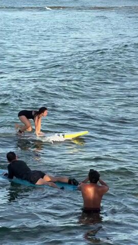 A woman wearing a black one piece swimsuit is surfing in a body of water. There are a few people in the water with her, also on surfboards. The woman starts in a kneeling position and then stands up as the board moves forward. She raises her arms to balance herself. The board comes to a stop, and the woman falls into the water.