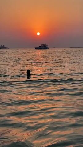 A person is swimming in the ocean at sunset. The sun is setting over the horizon, casting a warm orange glow across the water. A yacht is visible in the distance. The water is choppy, with small waves breaking on the shore. The person is partially submerged in the water, with their head and shoulders visible. They appear to be swimming towards the shore. The overall scene is peaceful and serene.