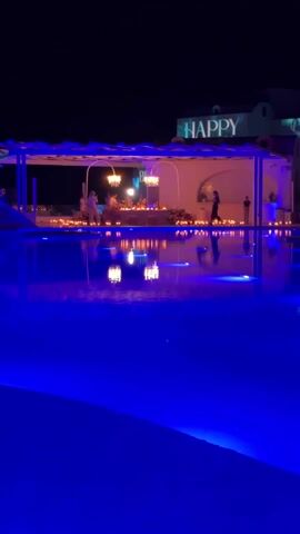 A nighttime shot displays a serene pool reflecting a candlelit pergola structure with "HAPPY" projected onto the rooftop. People are gathered within the structure, possibly celebrating a special occasion. The pool is lit with blue lights. A person walks alone on the deck beside the pool. The scene suggests a romantic and festive atmosphere.