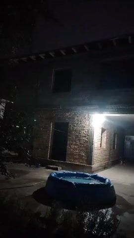 The video begins with a poorly lit, grainy shot of a building at night. The camera is shaky, and it's difficult to make out specific details. There appears to be an outdoor area in front of the building, possibly a courtyard or street, with some sort of blue object in the foreground. The building itself has windows and a doorway, and there are lights on inside. The camera then pans upwards, showing the facade of the building and then the dark sky. The remainder of the video is completely dark, making it impossible to see anything. It's possible the video was taken during a rainstorm, as there are some streaks that could be interpreted as rain.