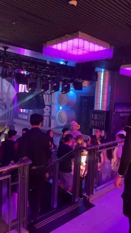 The video shows a crowded indoor space that appears to be a nightclub or a similar entertainment venue. The lighting is dim and predominantly purple, with spotlights and neon signs adding to the atmosphere. In the foreground, a railing and stairs are visible, with several people standing on them. One person on the stairs is holding a bottle and appears to be drinking. Further back, a group of people are gathered, including a person with blonde hair and a distinctive outfit who is standing out from the crowd. Behind this group, there is a large, futuristic-looking structure with circular elements and signage in what appears to be Korean characters. Other individuals are scattered throughout the scene, some interacting, others observing. The overall impression is one of a lively and energetic gathering.