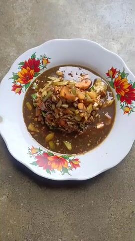A white ceramic plate with a floral pattern of red and yellow flowers is centered in the frame. The plate contains a serving of food, which appears to be a stew or soup with various chopped vegetables such as carrots, tomatoes, and green onions, mixed with some shredded meat or fish, all in a dark brown broth. The plate is placed on a textured, greyish-brown surface, possibly concrete or stone, with visible grain and slight imperfections. The lighting is natural and diffused, casting subtle shadows. The camera angle is directly overhead, looking down at the plate of food. No people or other objects are visible in the frame. The video consists of a static shot of the food.