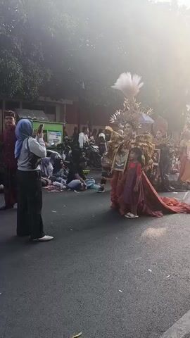 The video shows a street scene with people gathered, possibly for an event or parade. In the foreground, a person wearing a headscarf and black pants is taking a photo with a smartphone. To their right, two individuals are dressed in elaborate, ornate costumes. One person wears a predominantly red costume with a long, flowing train and a large, feathered headdress. Beside them, another person is also in costume, with a golden, intricate headdress. A young child in a red dress stands near the costumed individuals, holding a framed picture. In the background, more people are visible, some sitting on the ground and others standing, with what appear to be vendor stalls or displays. The environment appears to be outdoors on a paved street, with trees and buildings visible in the distance. The lighting suggests it is daytime, with the sun creating a hazy or slightly overexposed effect.