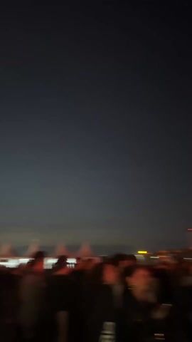 The video shows a crowd of people at night, with red and orange lights illuminating the foreground and a dark sky above. The camera movement is shaky and suggests that the videographer is part of the crowd.  The lights are indistinct, but may be coming from structures or vehicles in the distance. The focus is soft and details are blurred.
